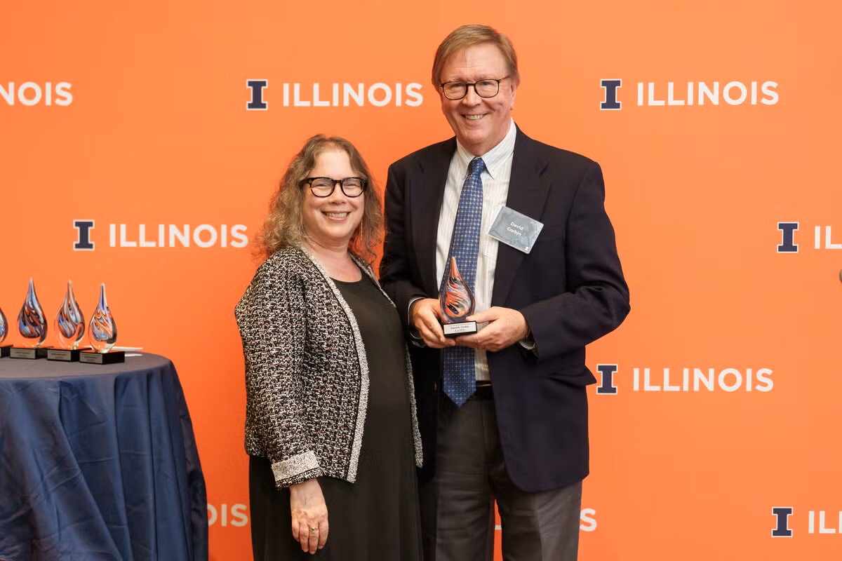woman stands next to a man holding a trophy in front of an orange background with the text "Illinois"